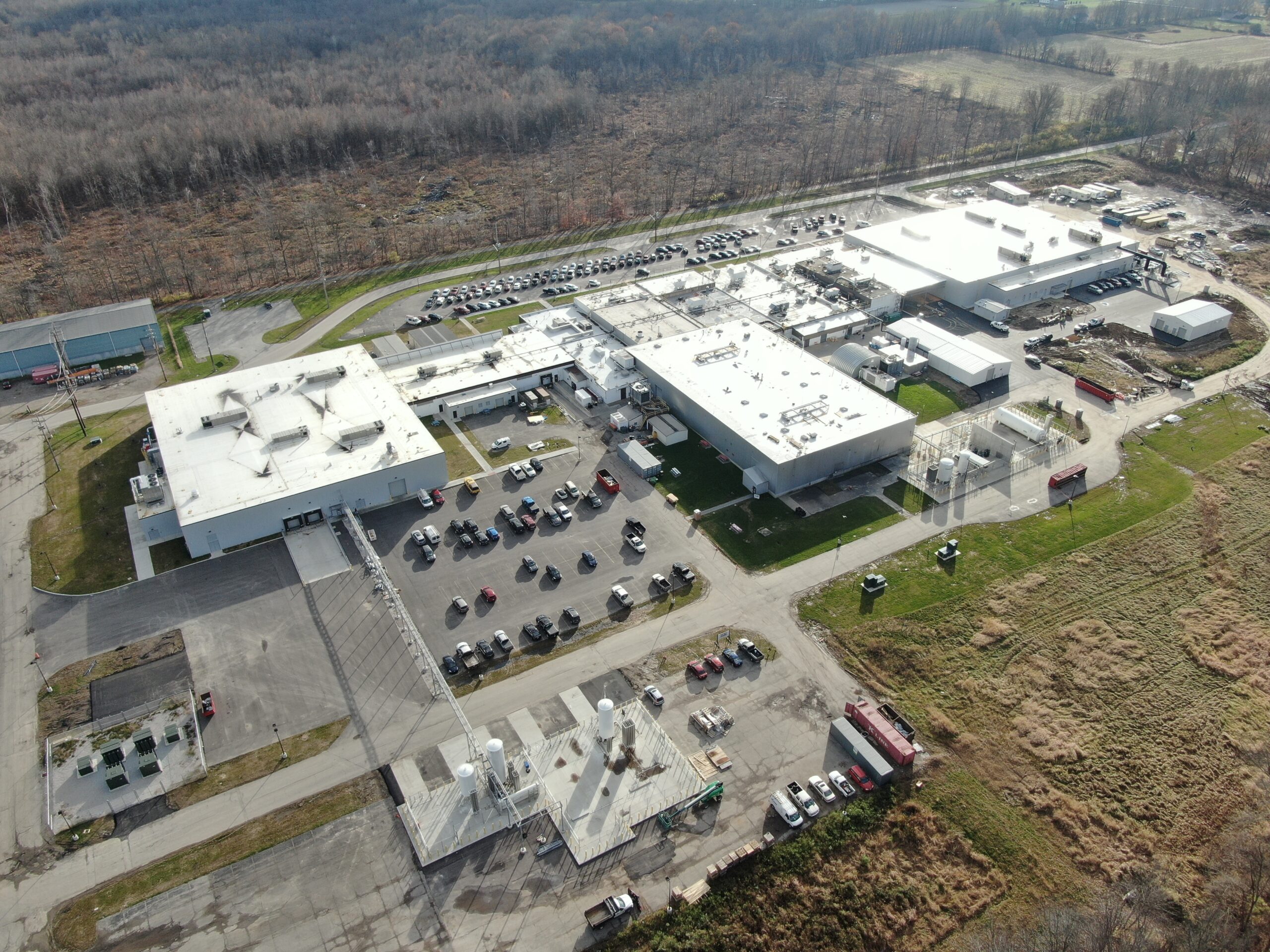 Aerial view of a large industrial complex with flat-roofed buildings, paved parking lots, and several transport trucks and cars parked throughout the facility.