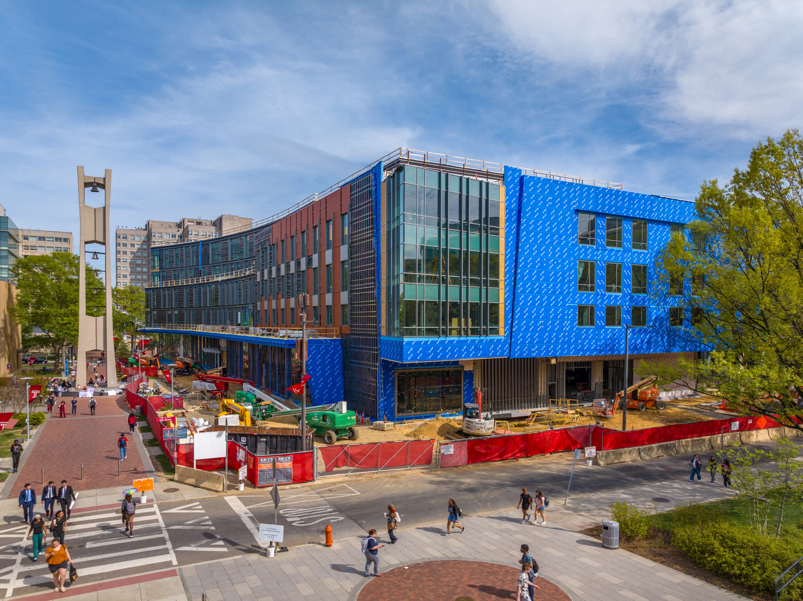 A multi-story building under construction, wrapped in blue weather-resistant material, located on a university campus with students walking on a nearby plaza.