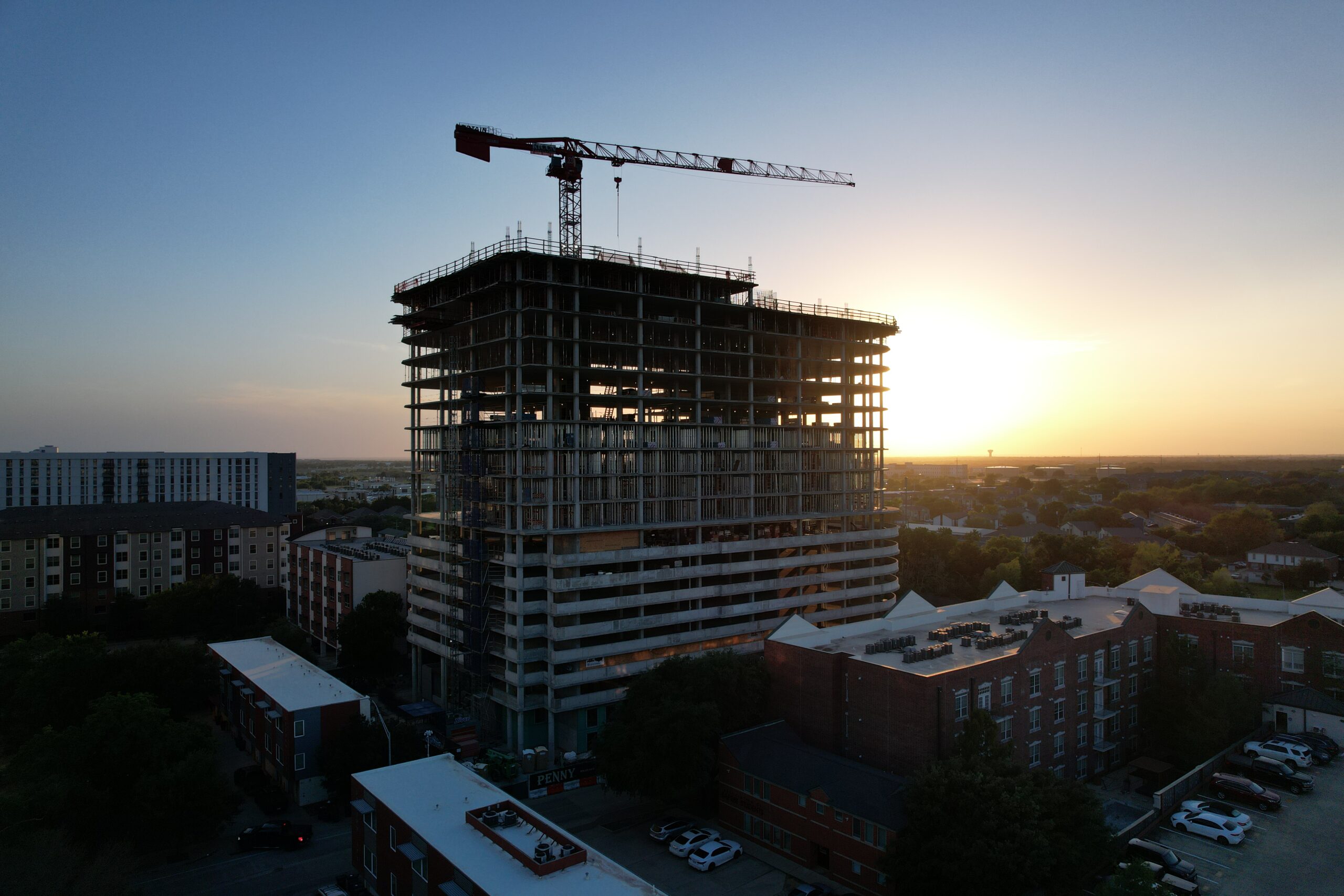 An aerial view of a high-rise building under construction, silhouetted against a setting sun with a tower crane on top.