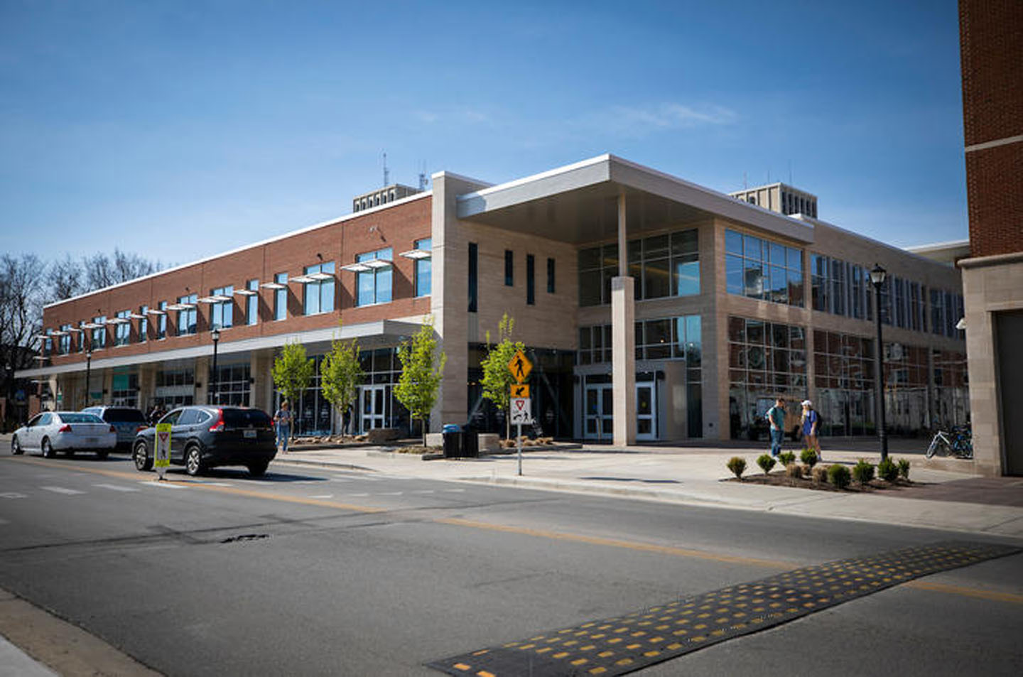 Modern brick and glass building on a street corner with a pedestrian crossing, cars, and people walking.