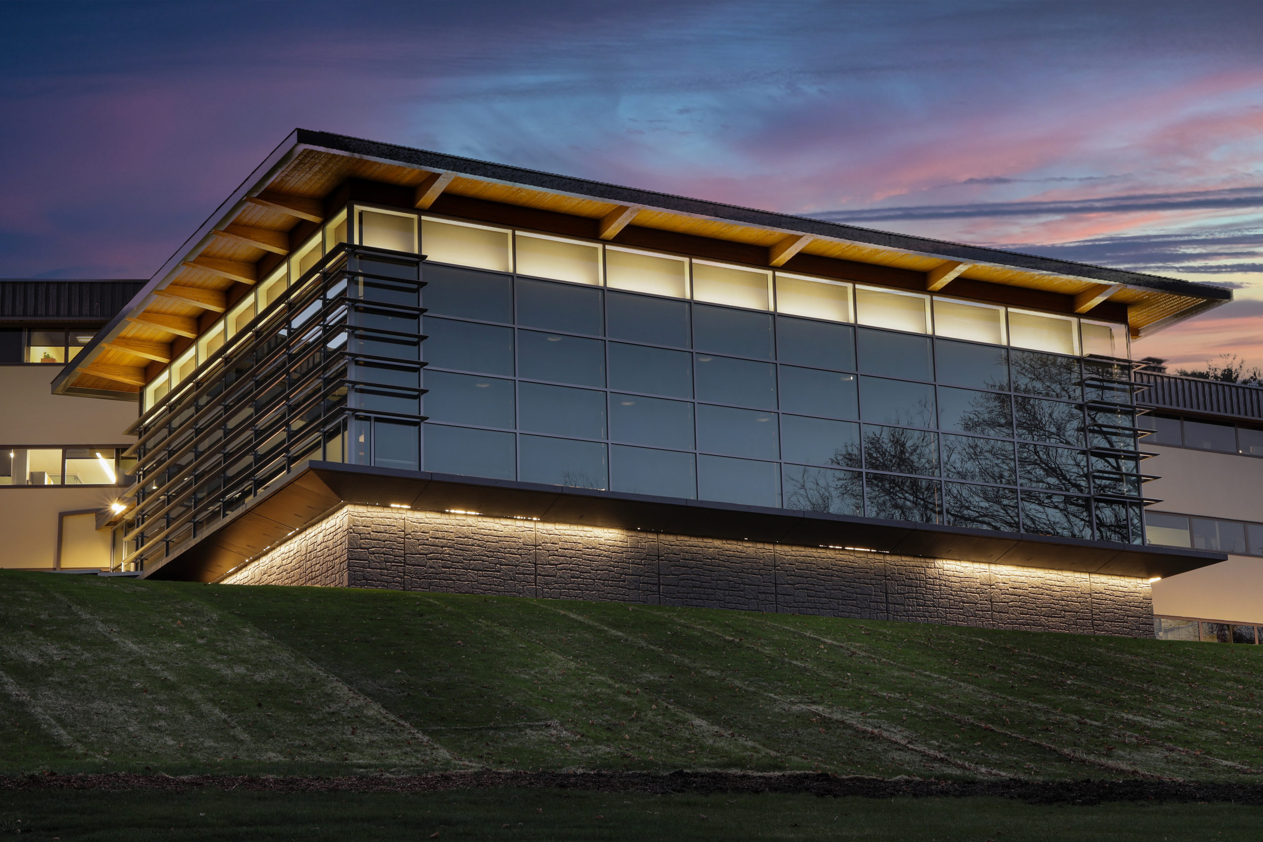 Modern office building with a glass facade and wooden roof overhang, set on a grassy hill at twilight.