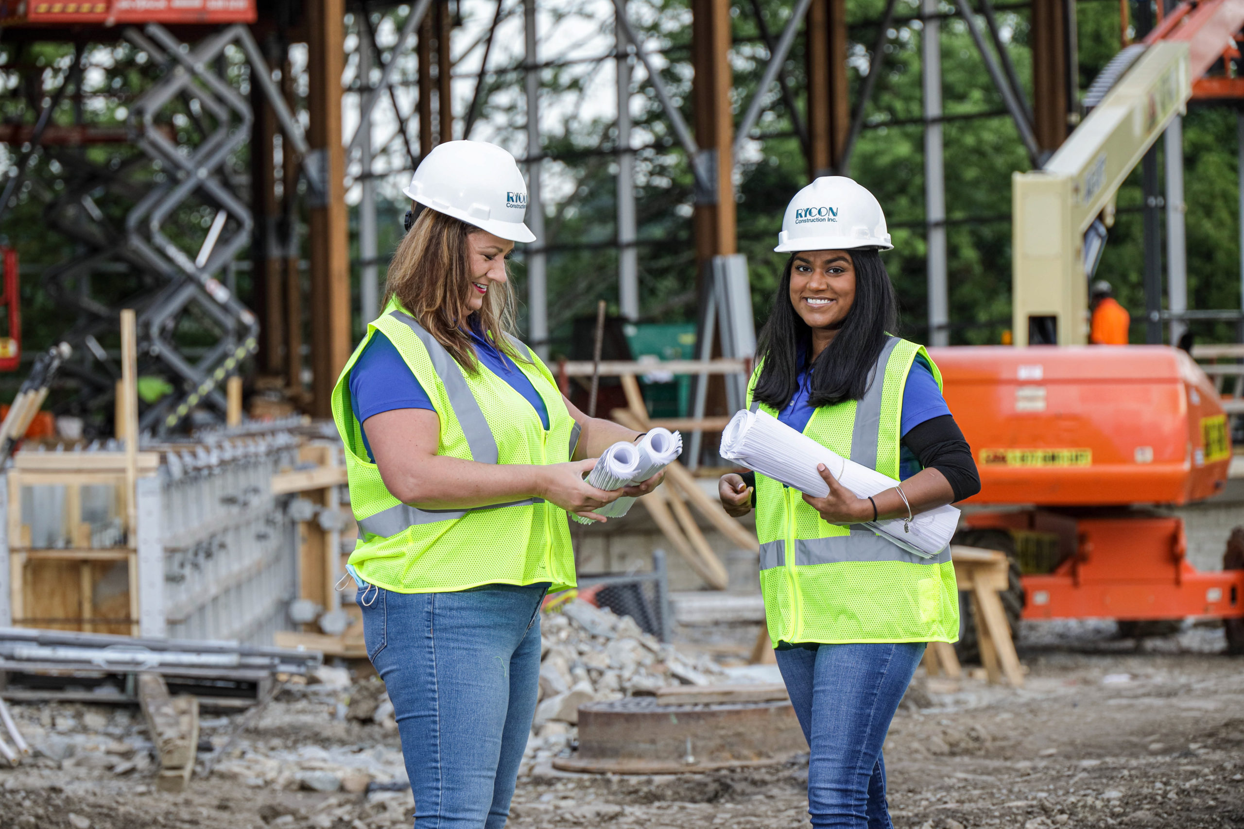 Two construction project managers in hard hats and high-visibility vests standing at a job site while reviewing building plans.
