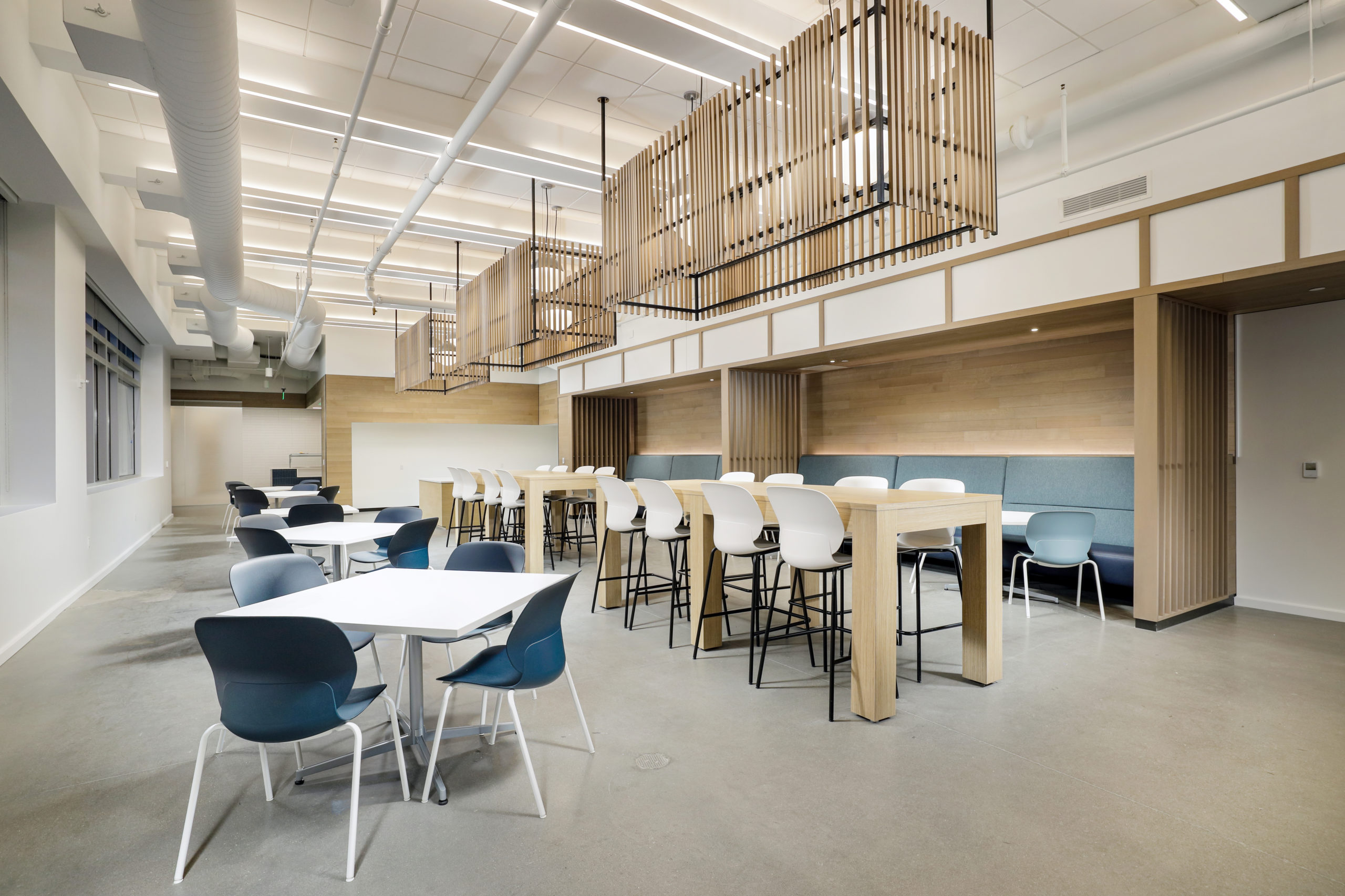Modern office break room with white square tables, blue chairs, and a long wooden bar-height counter with matching stools under slatted ceiling light fixtures.