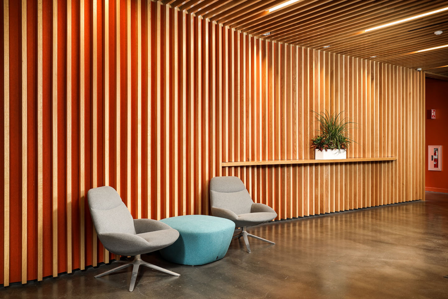 Two grey chairs and a teal ottoman in a lobby with slatted wood walls and a built-in shelf holding a potted plant.
