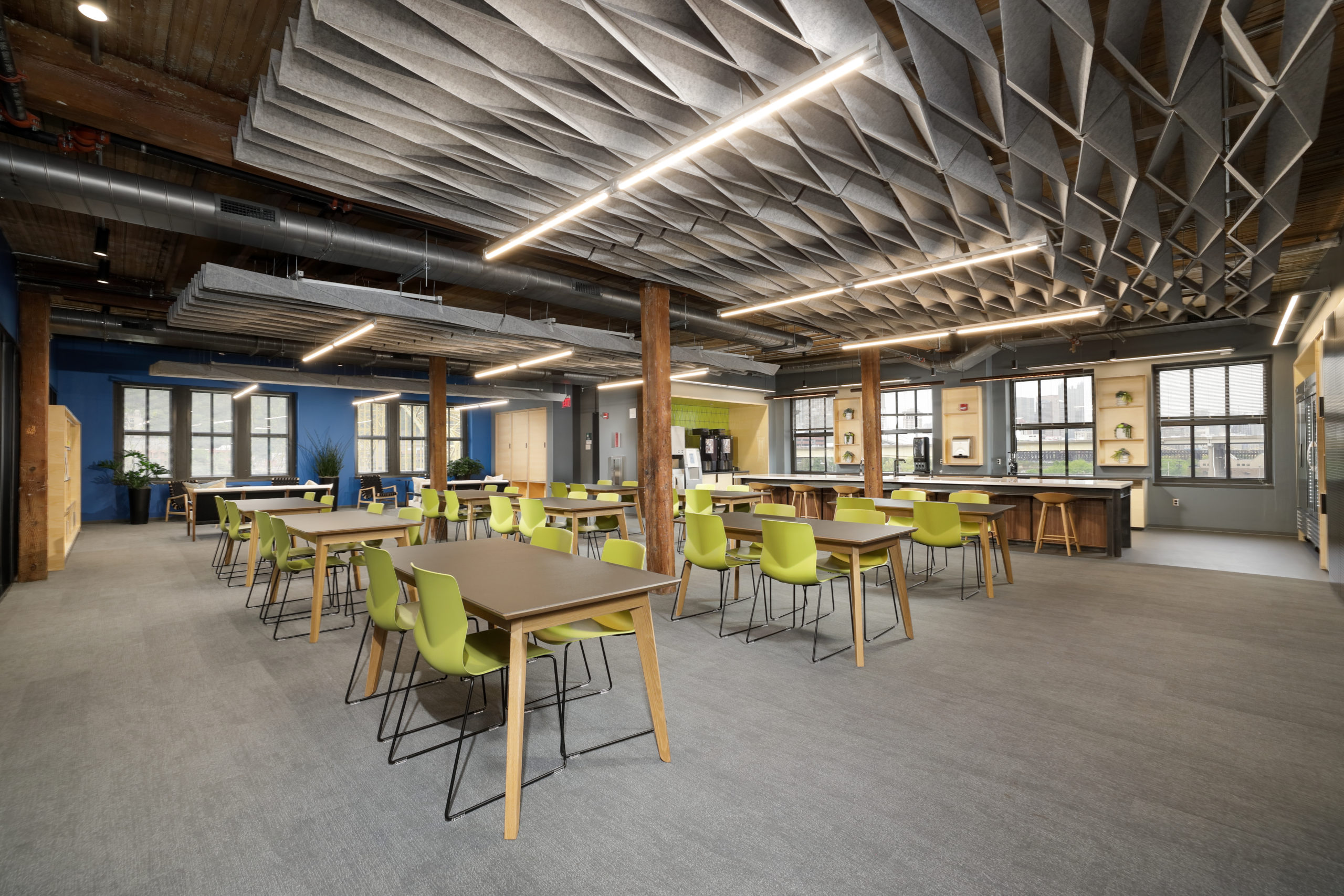 A modern open-plan office breakroom with rows of tables, lime green chairs, and a geometric acoustic ceiling.