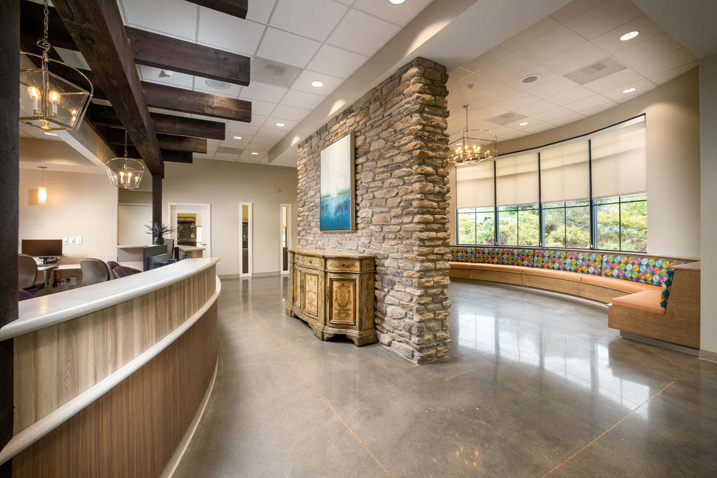 Reception area with a curved wooden desk, stone accent wall featuring a painting and wood sideboard, and a long curved bench with colorful cushions under windows.