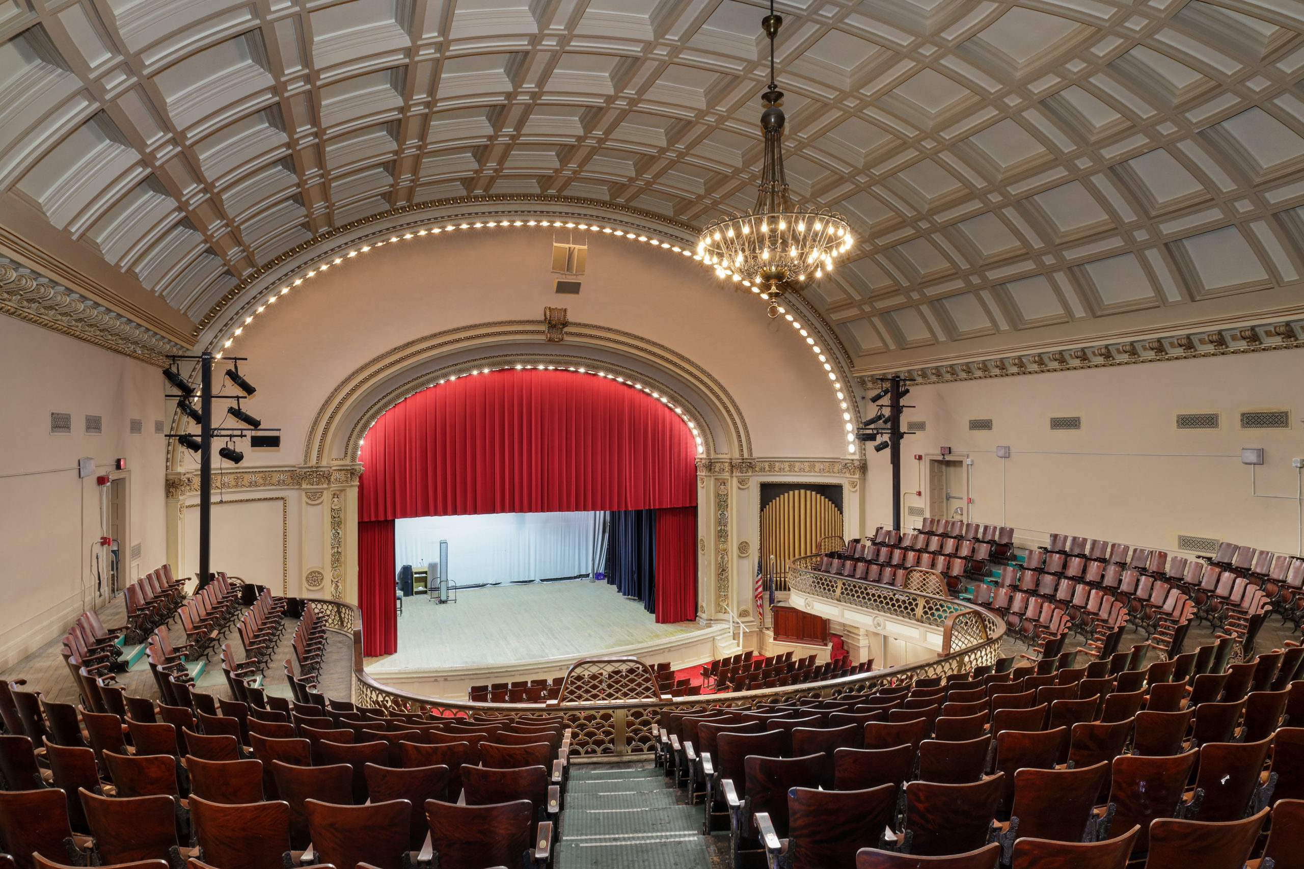 Interior of a historic theater with rows of wooden seats facing a stage framed by a large red curtain.