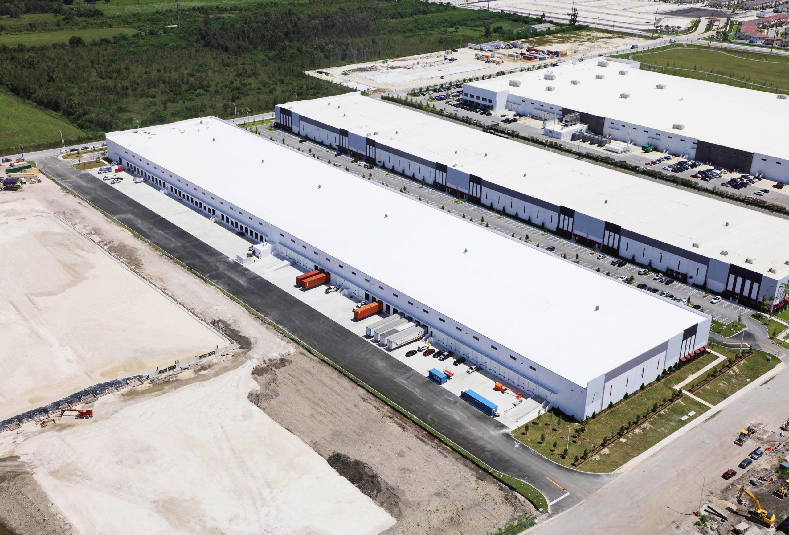 Aerial view of large industrial warehouse buildings with flat white roofs, shipping bays, and adjacent paved parking lots.