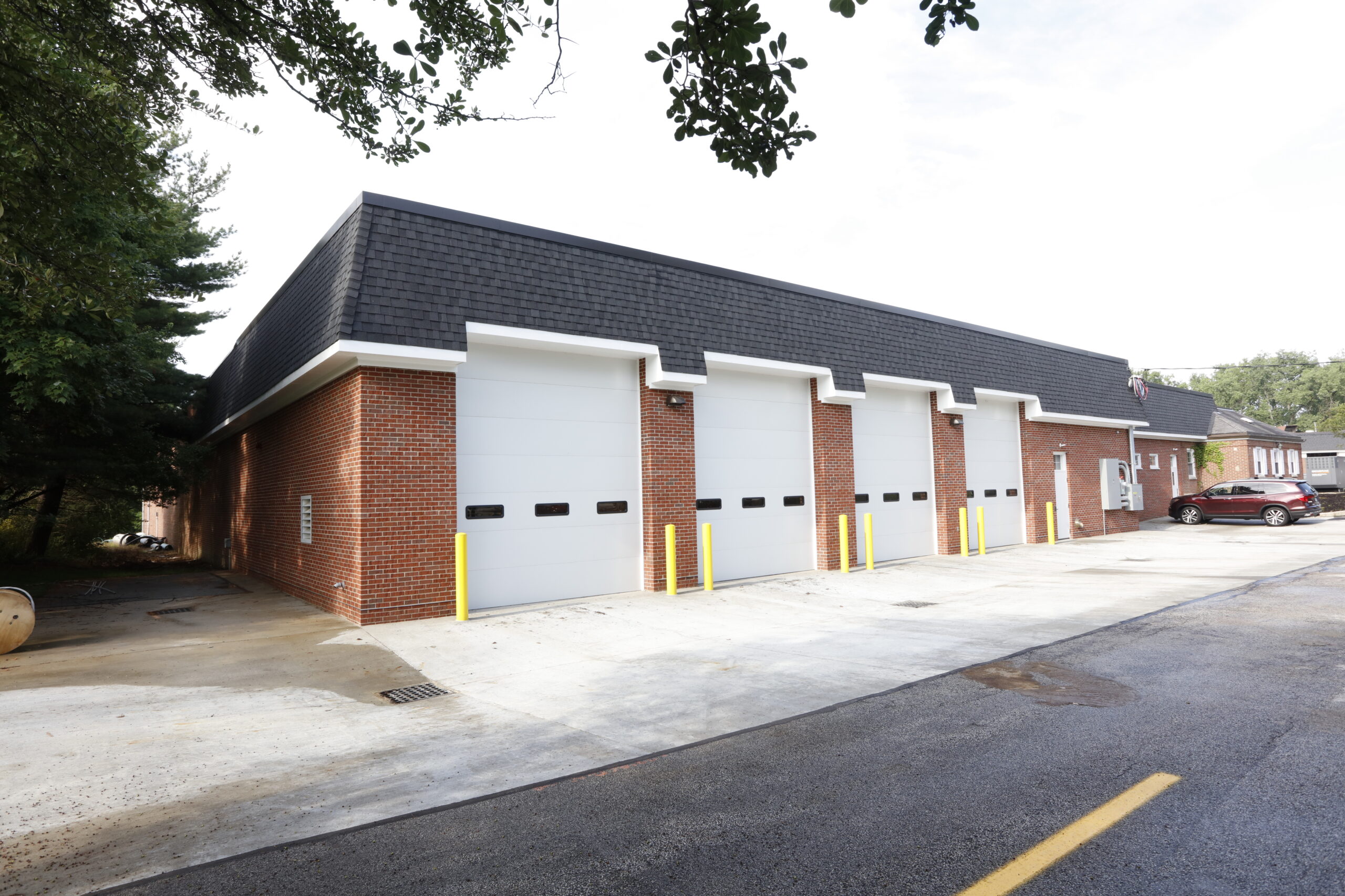 A single-story red brick building featuring four white garage bay doors and a dark shingled mansard roof.