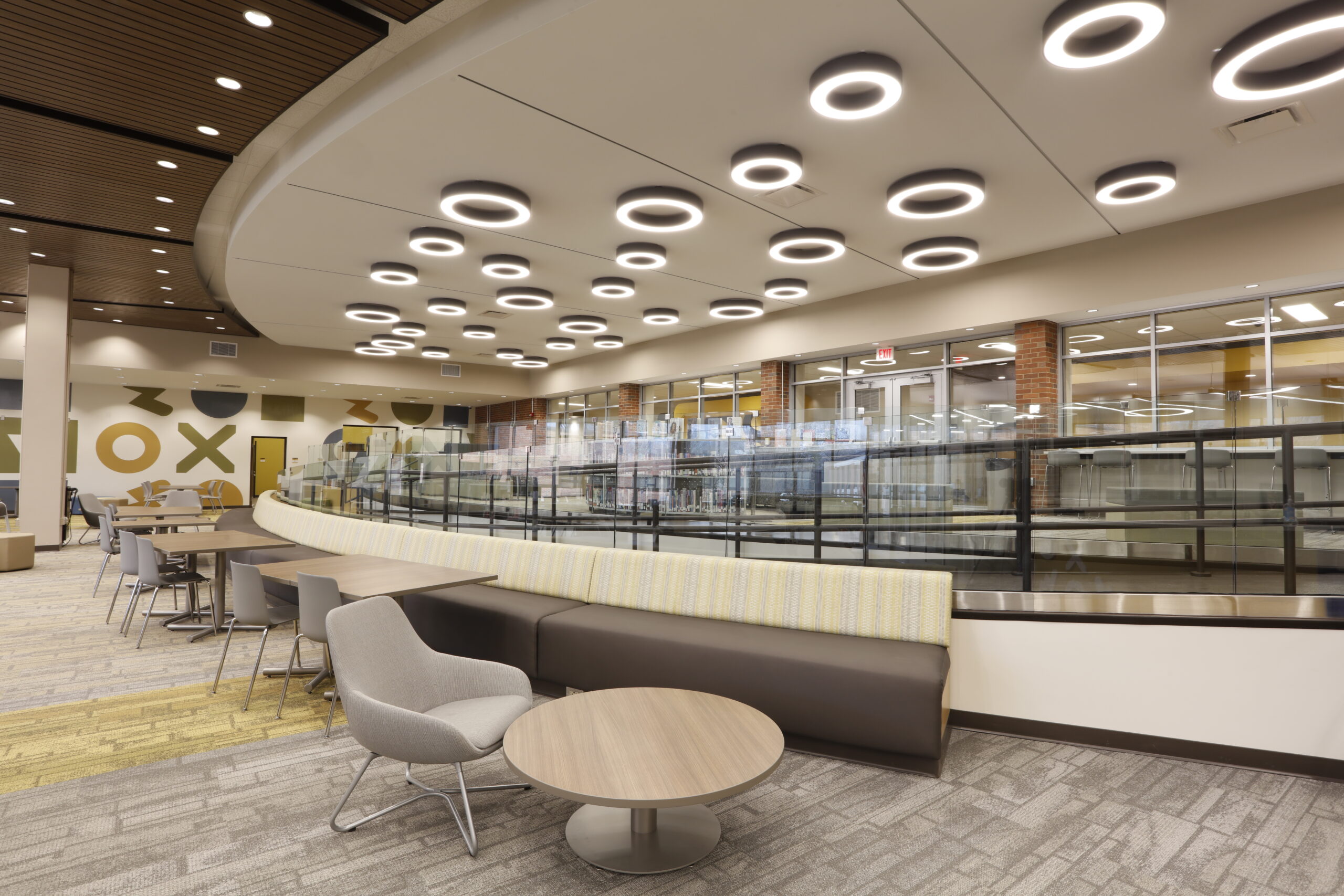 Modern library lounge featuring a long, curved banquette seating area with small round tables and individual chairs, under a ceiling with numerous circular LED lights.