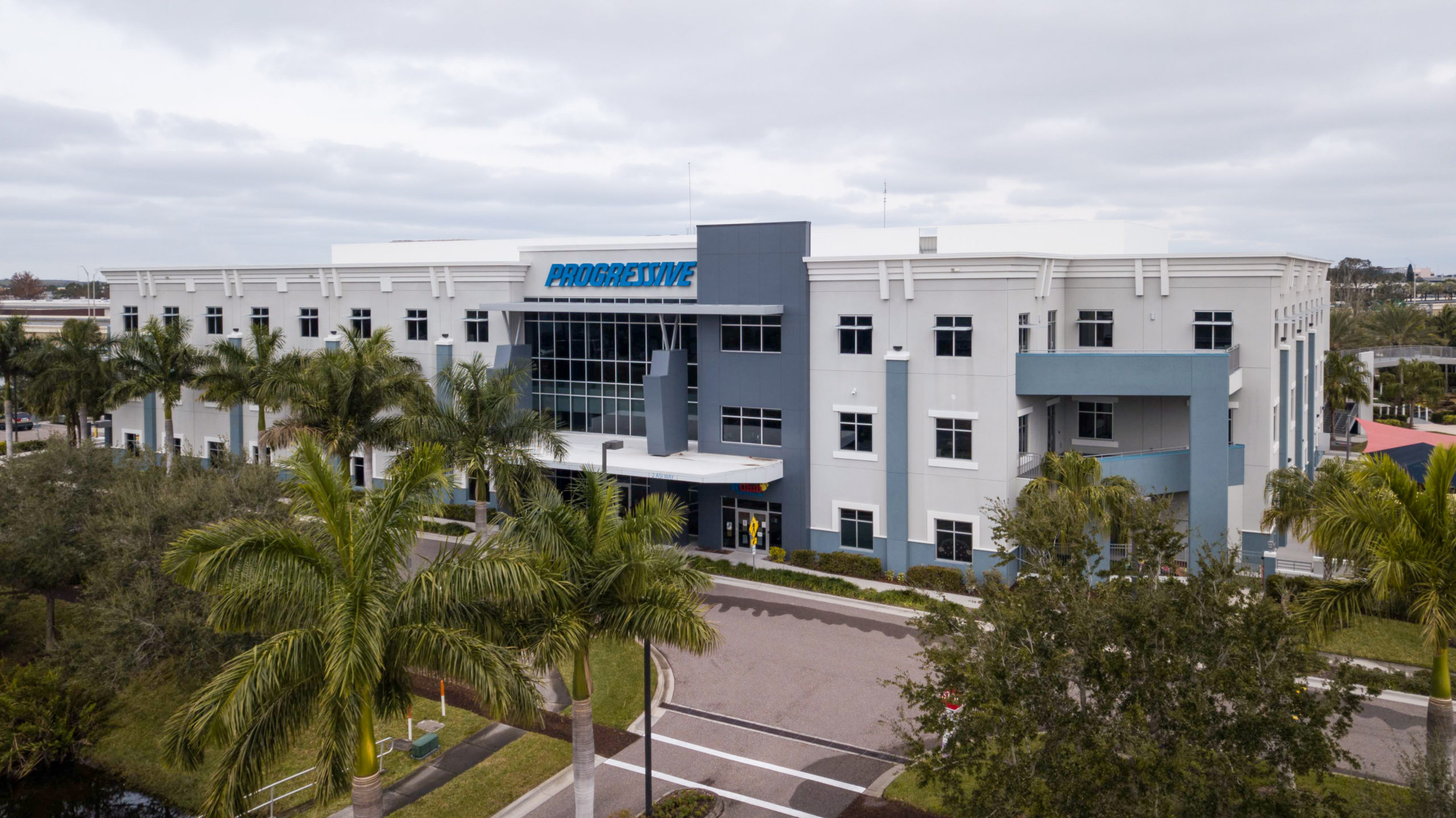 Aerial view of a multi-story Progressive office building surrounded by palm trees and a paved driveway under a cloudy sky.