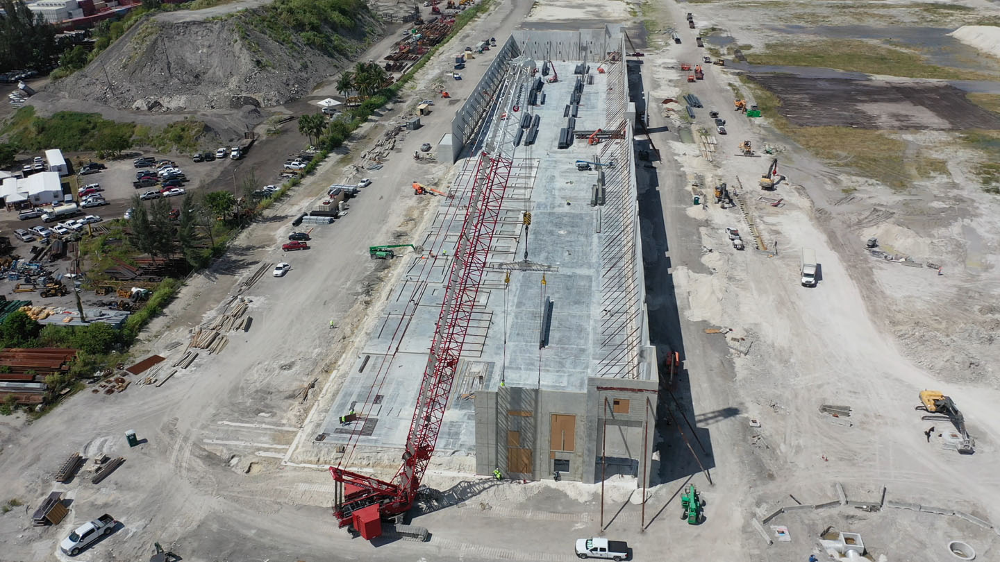 Aerial view of a large concrete building under construction with a red crane lifting a steel beam.