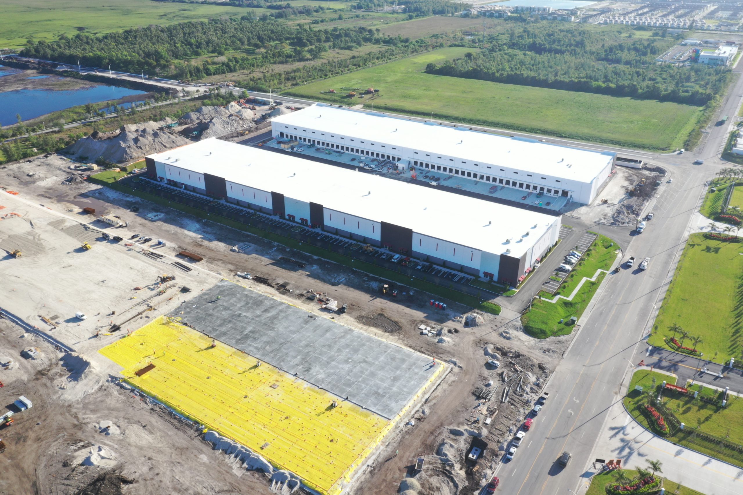 Aerial view of two large industrial warehouse buildings surrounded by construction zones, parking areas, and a nearby road.