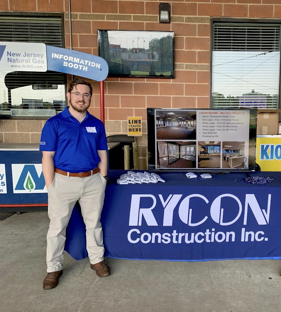 A man in a blue polo shirt stands in front of a Rycon Construction Inc. information table.