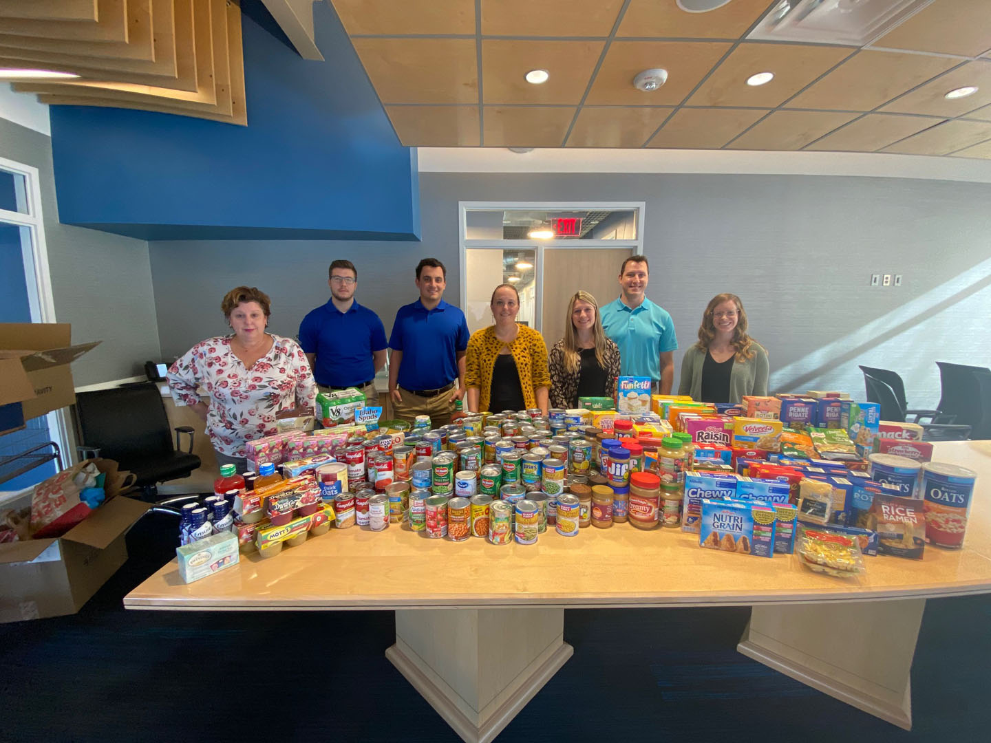 Six people standing behind a large conference table covered with a variety of canned and boxed food donations.