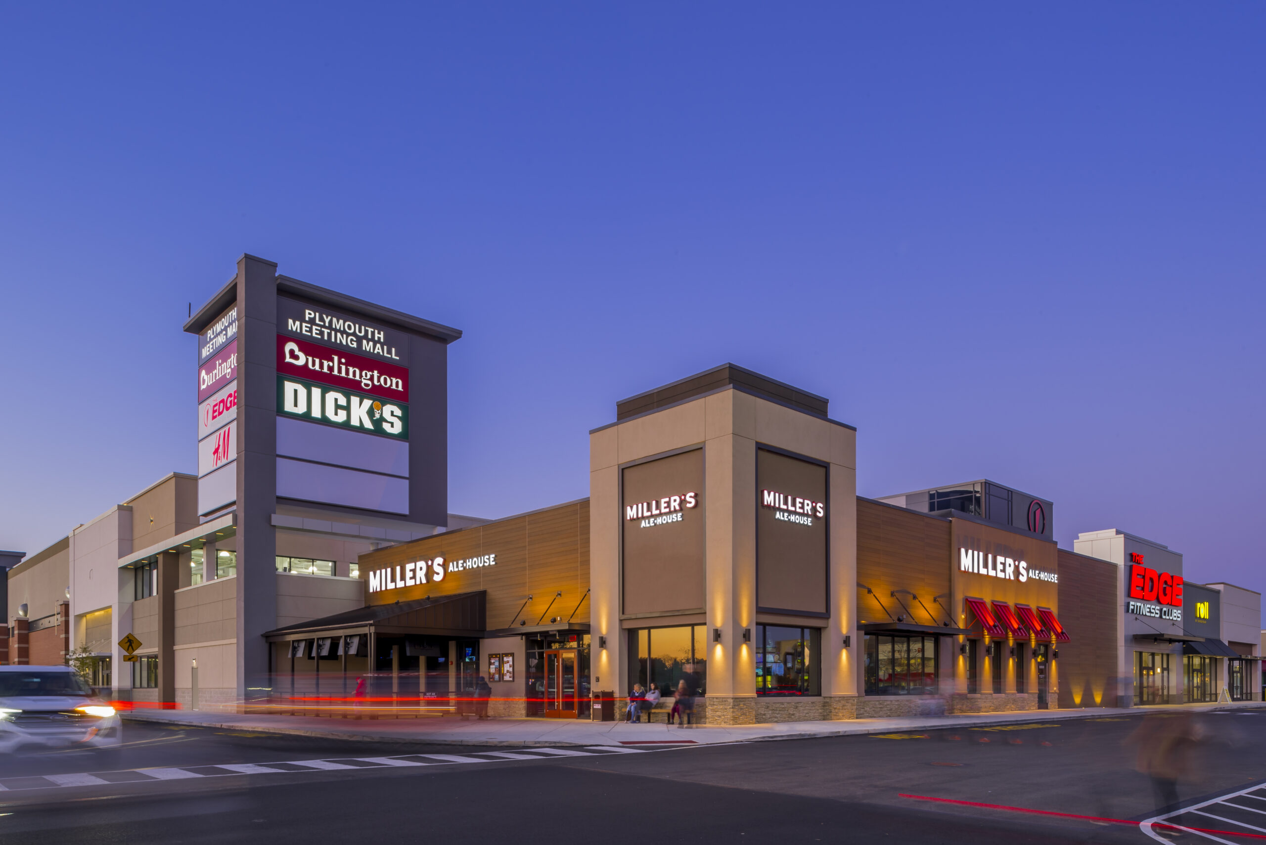 Exterior of Plymouth Meeting Mall at dusk, featuring a tall pylon sign with Burlington and Dick’s branding, and a corner Miller’s Ale House restaurant.
