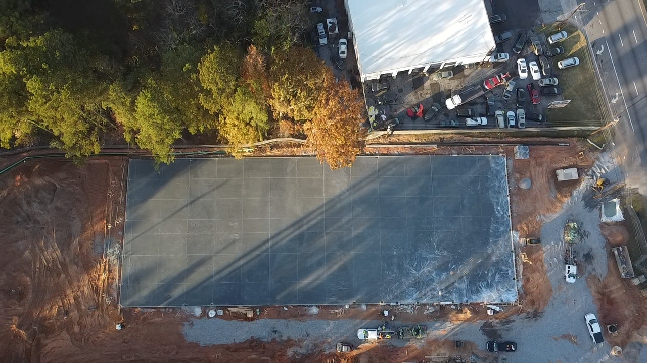 Aerial view of a rectangular concrete foundation slab at a construction site next to a wooded area and a parking lot.