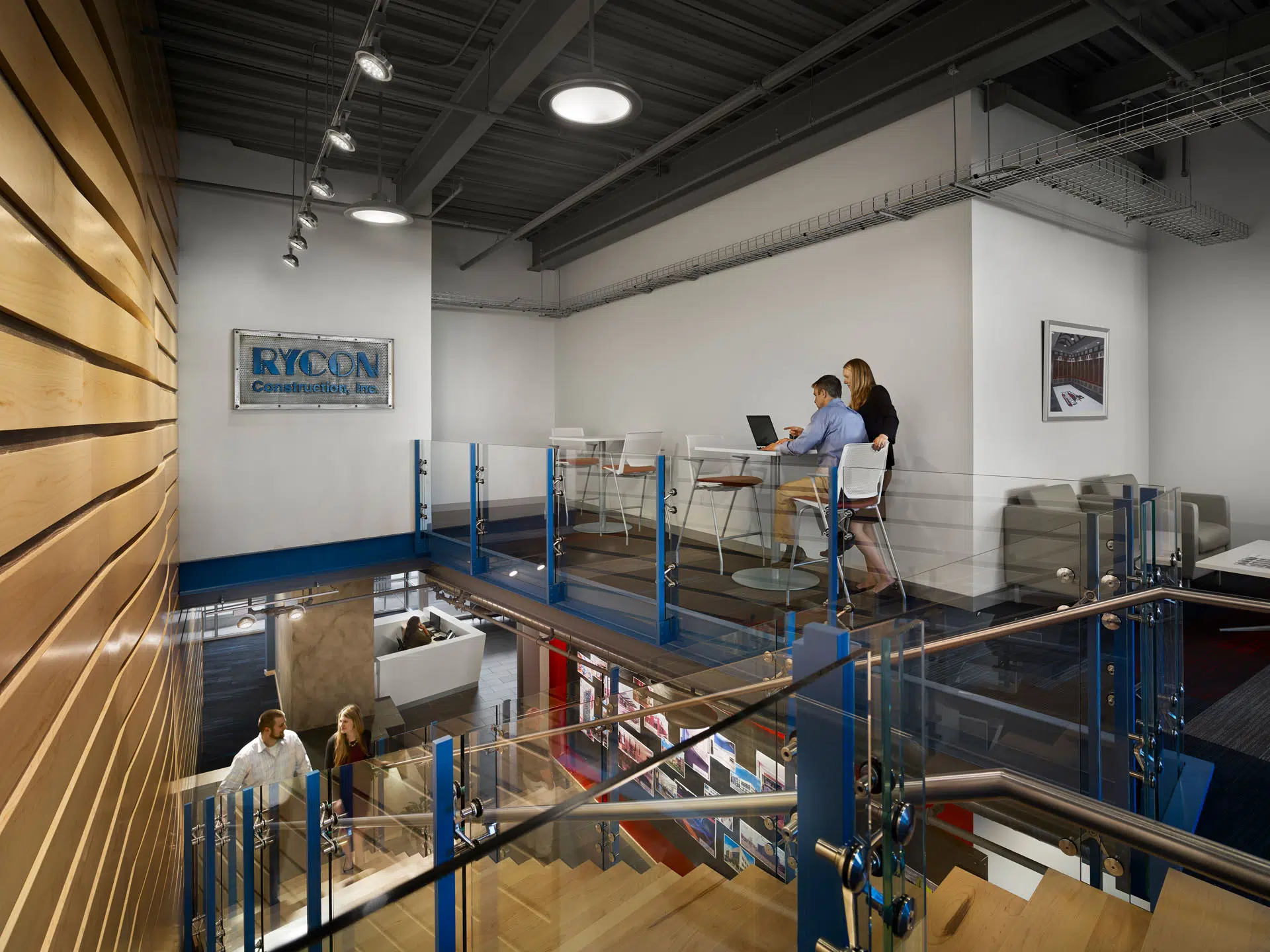 An open-plan office space with a Rycon Construction sign on the wall, showing people working at a lofted desk area and walking up a wooden staircase.