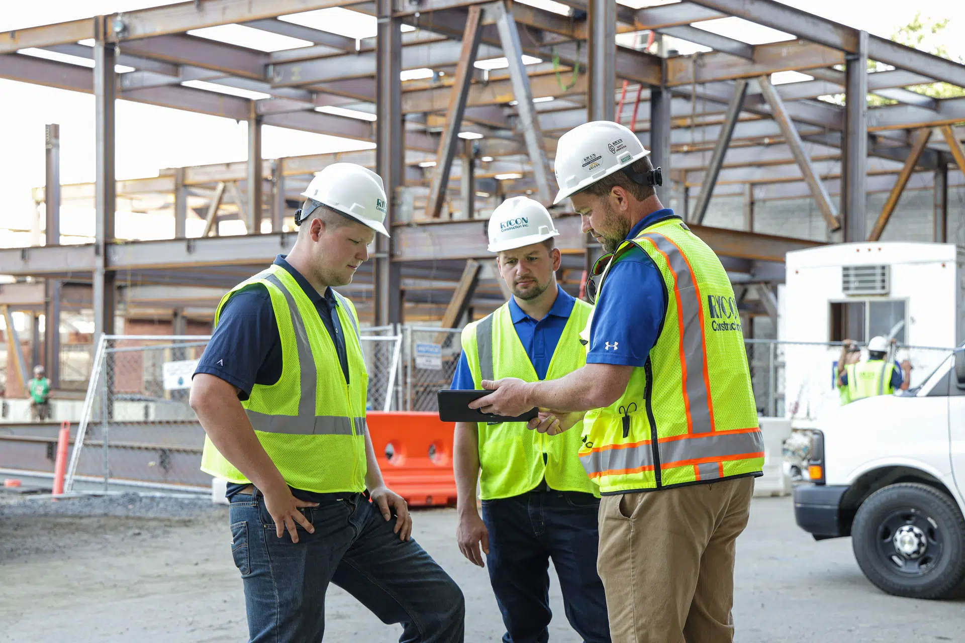 Three construction workers in hard hats and high-visibility vests look at a tablet at a building site.