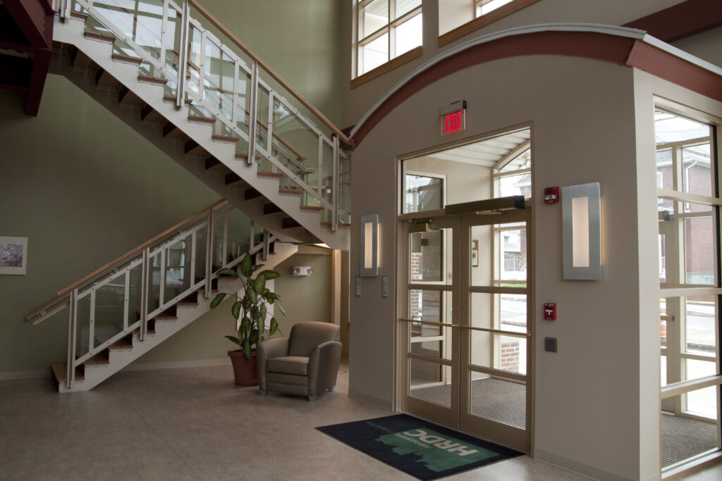 Interior lobby with a wooden staircase, a single armchair, and glass entry doors marked with an exit sign.