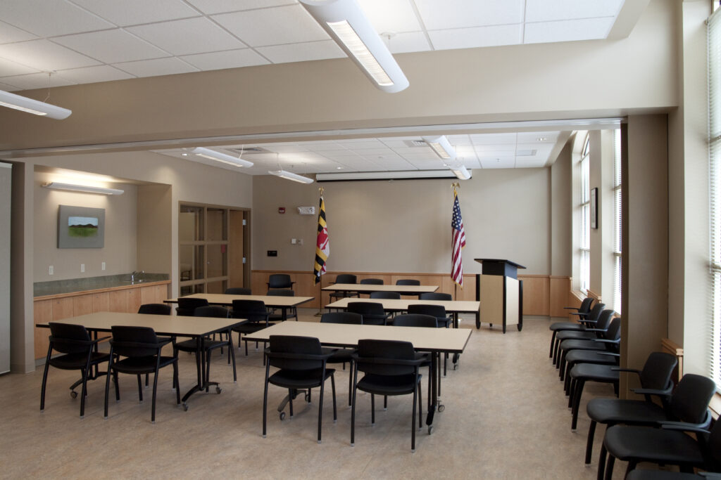 A classroom or meeting room with tables and chairs arranged in rows, featuring a podium and the flags of Maryland and the United States.