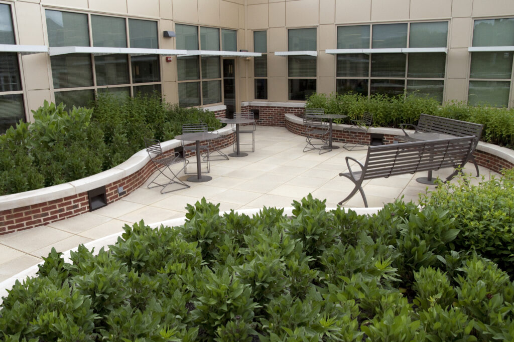 Outdoor patio with tables, chairs, and a bench surrounded by brick planters and green shrubs.
