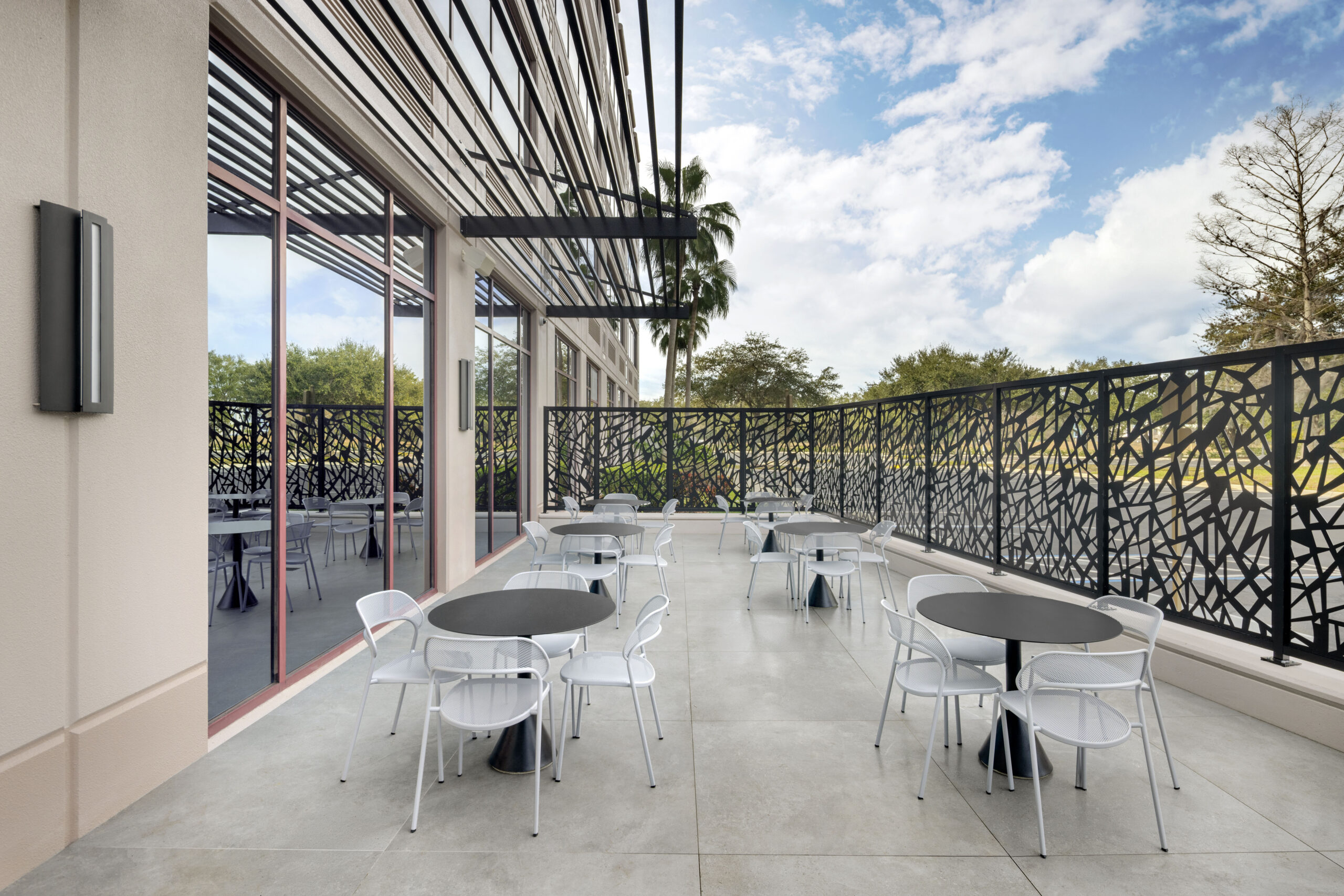 Outdoor patio with multiple small round tables and white metal chairs, enclosed by a decorative black metal fence.
