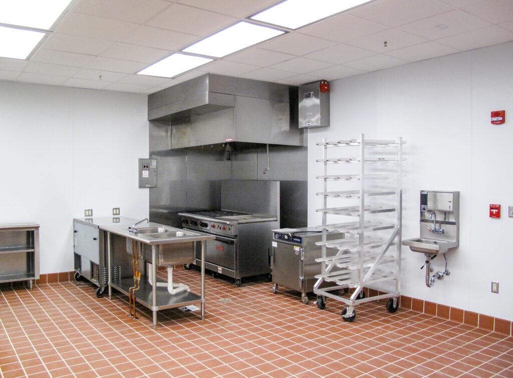 A commercial kitchen with stainless steel counters, a sink, a range under a ventilation hood, a rolling metal rack, and a wall-mounted hand-washing sink on a red tiled floor.
