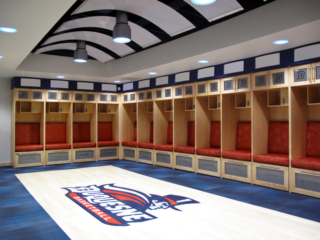 A sports locker room with wooden stalls, red cushioned seats, and the Duquesne Basketball logo on the floor.