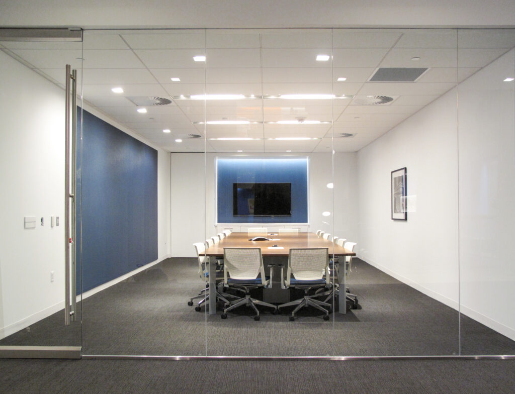 A glass-walled conference room with a long table, ergonomic chairs, and a wall-mounted television.