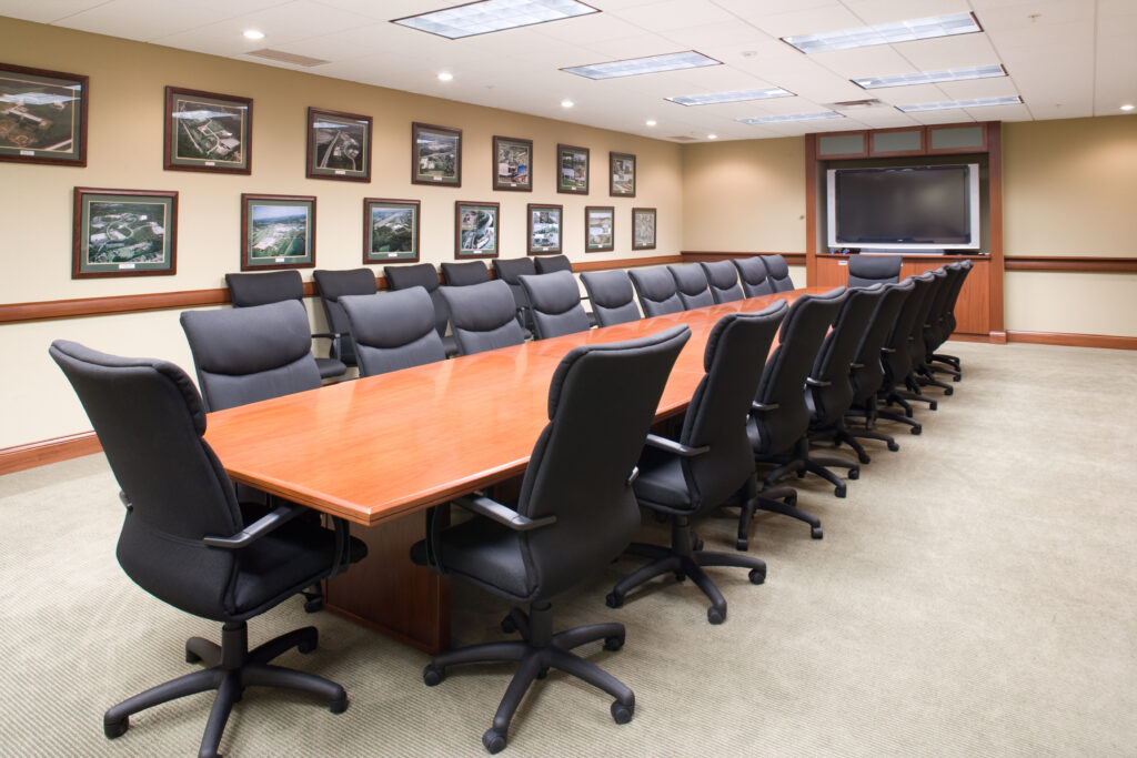 A conference room with a long wooden table surrounded by black office chairs, and framed photos of buildings hanging on the tan walls.