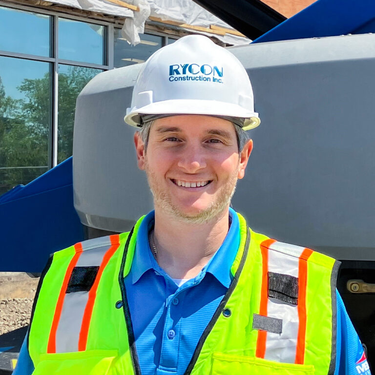 A smiling construction worker wearing a white Rycon Construction hard hat and a yellow high-visibility safety vest, standing in front of heavy machinery.