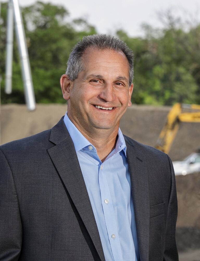 A smiling man in a suit jacket and light blue dress shirt, standing at a construction site.