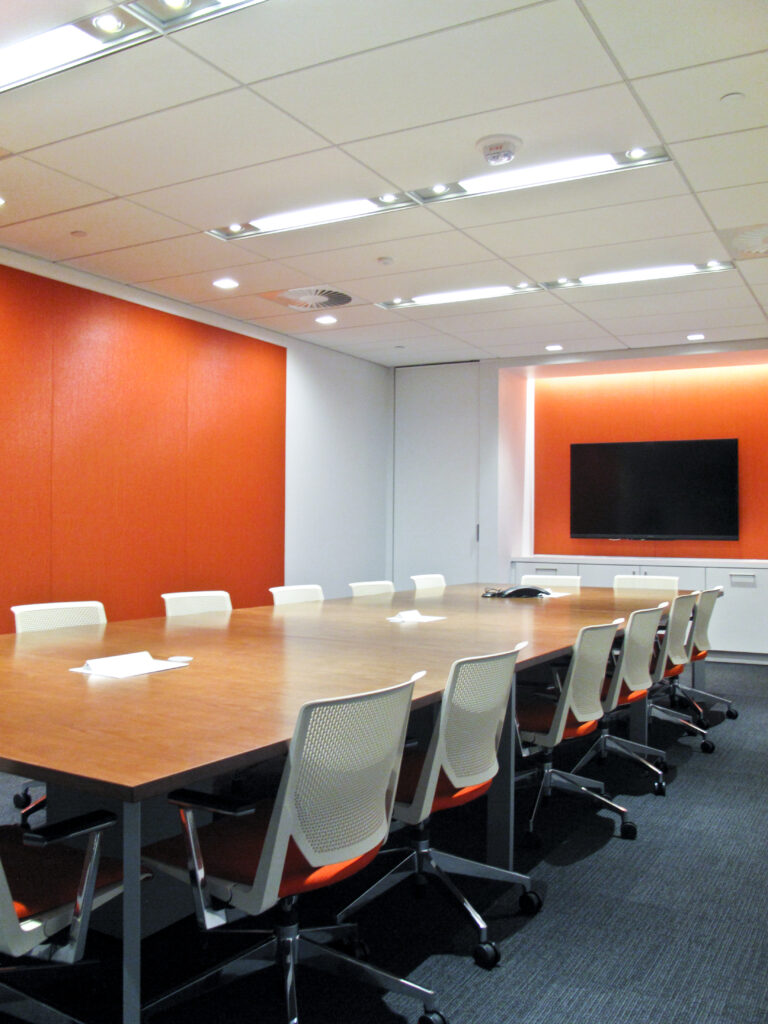 A conference room with a long wood-topped table, white office chairs, orange accent walls, and a wall-mounted television.