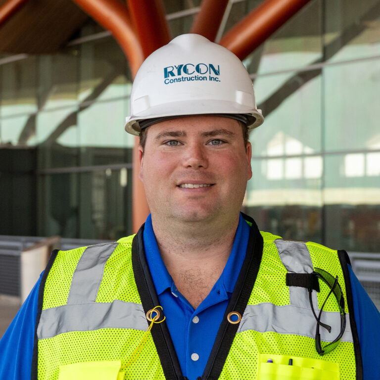A construction worker wearing a white Rycon Construction hard hat and a high-visibility safety vest over a blue polo shirt.