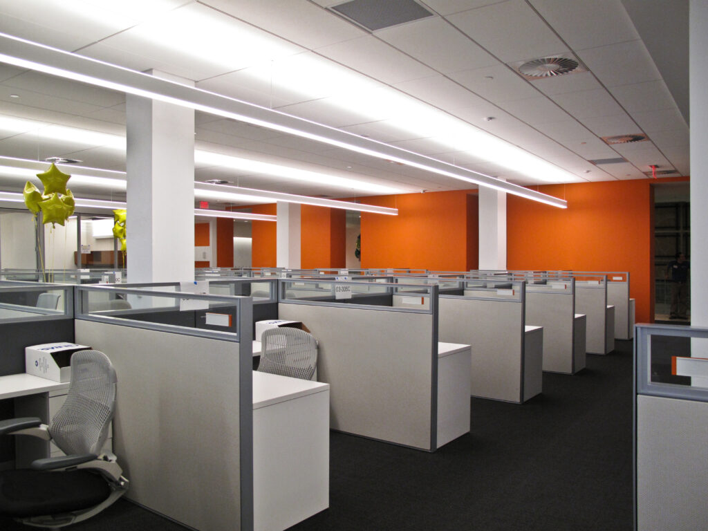 Rows of empty office cubicles with white desks and gray partitions in an office with orange walls.