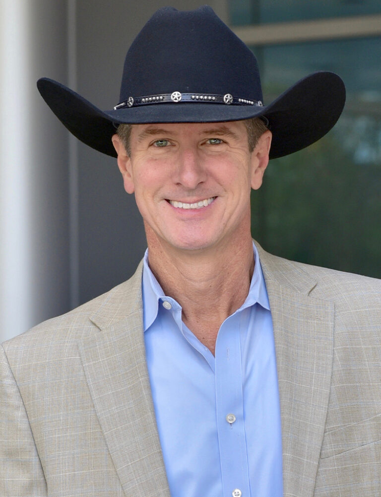 A man in a black cowboy hat, blue button-down shirt, and tan blazer smiling at the camera.