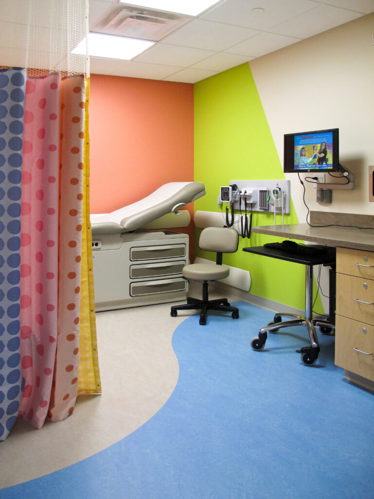 A pediatric exam room with a medical exam table, swivel stool, wall-mounted screen, and colorful, patterned privacy curtains.