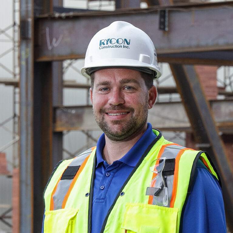 A smiling construction worker wearing a hard hat with the Rycon Construction Inc. logo and a high-visibility safety vest at a construction site.