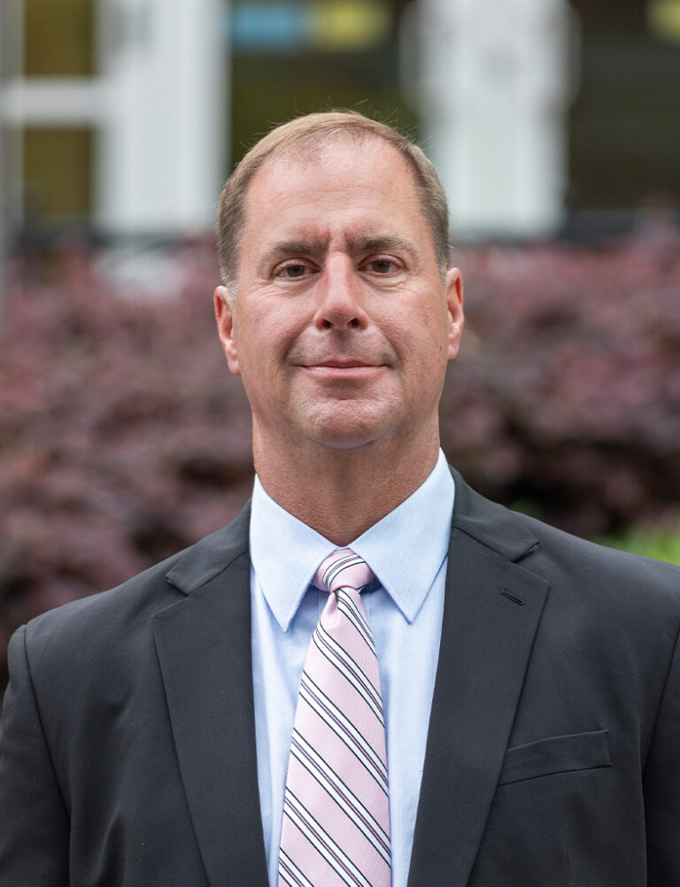 A middle-aged man in a black suit, light blue collared shirt, and pink striped tie.