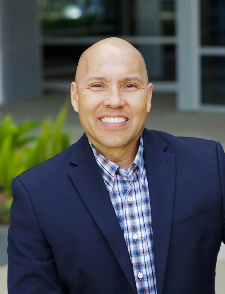 A smiling, bald man wearing a navy blue blazer over a plaid button-down shirt.