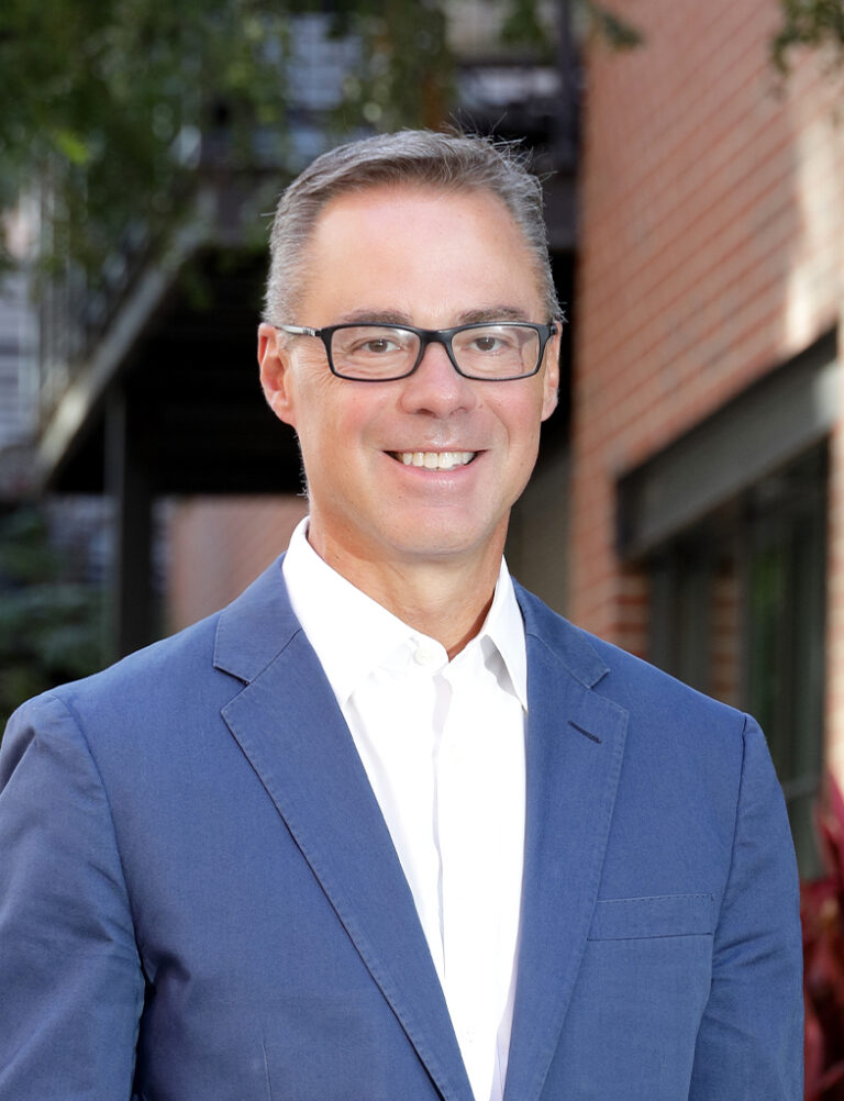 Headshot of a smiling man wearing glasses, a white collared shirt, and a blue blazer.