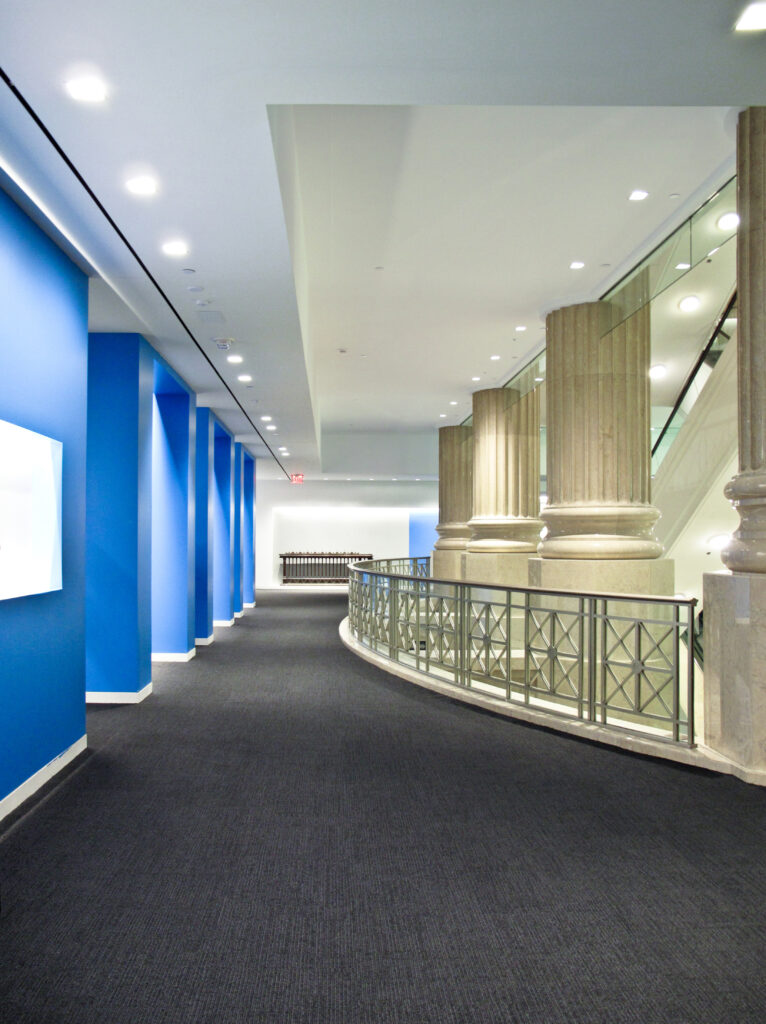 A curved hallway with blue accent walls on one side and a metal railing beside large stone columns on the other.