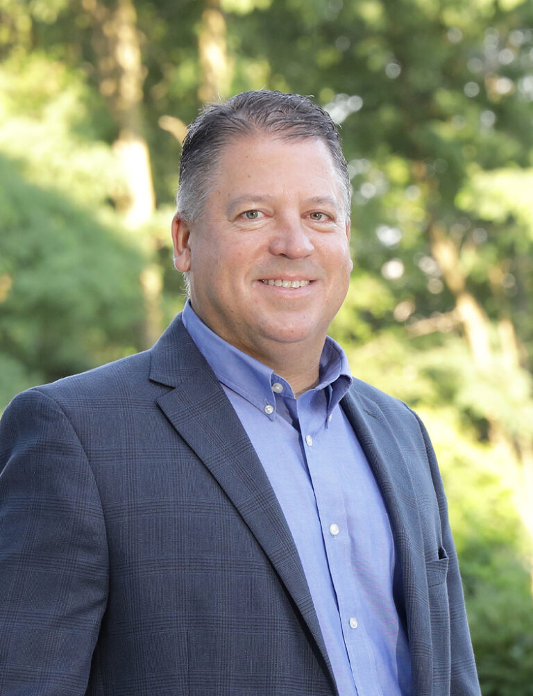 A middle-aged man smiling, wearing a blue dress shirt and a dark blazer.
