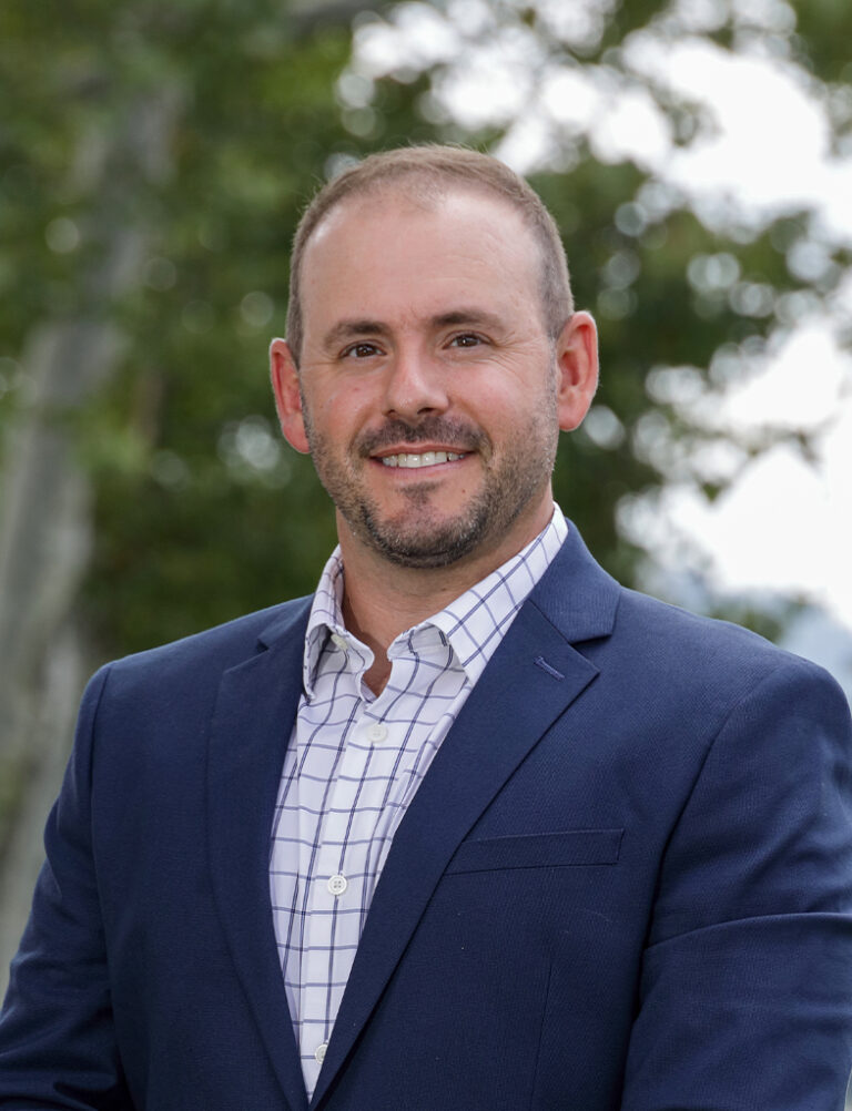 A man with short brown hair and stubble wearing a navy blue blazer and a white checkered dress shirt.