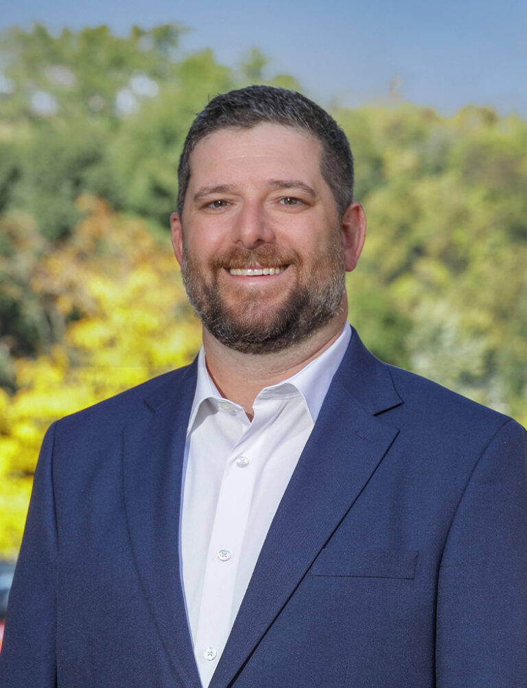 A man with a beard wearing a white button-down shirt and a navy blue blazer.