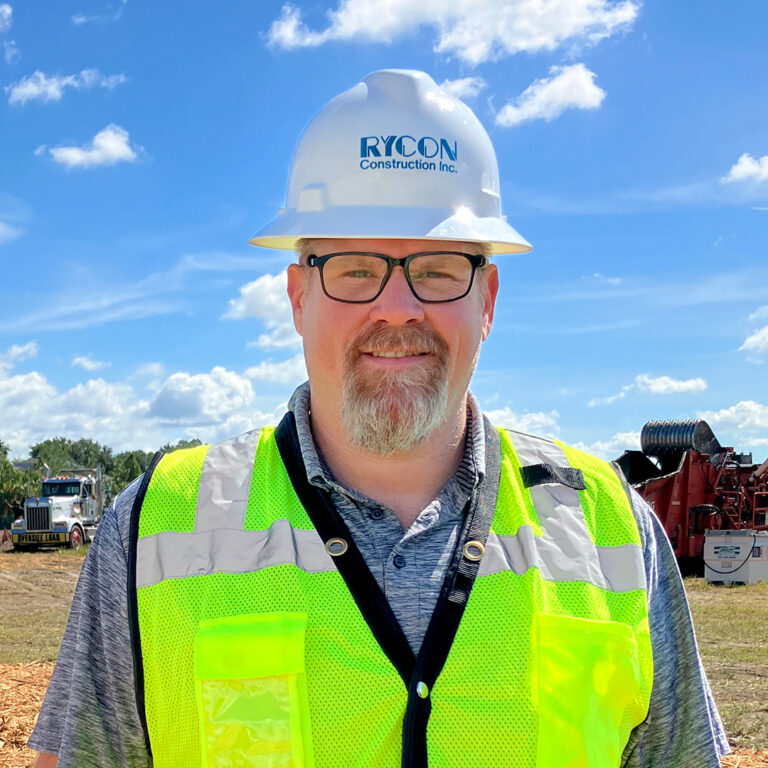 A man wearing glasses, a Rycon Construction hard hat, and a high-visibility yellow safety vest stands at a construction site.