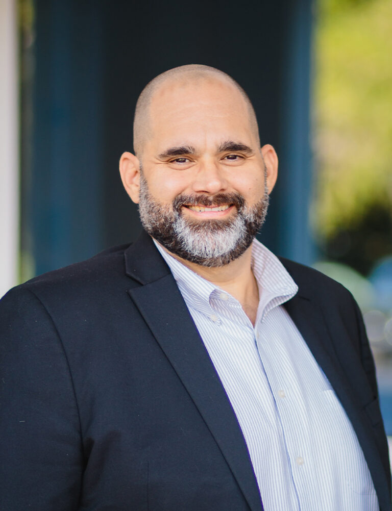 A smiling man with a shaved head and a beard, wearing a black blazer and a striped collared shirt.