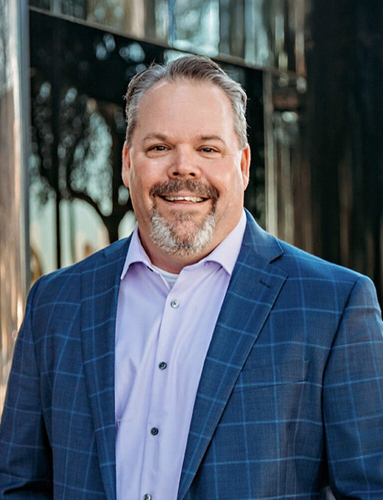 A smiling man with a goatee wearing a light purple button-down shirt and a blue plaid blazer.