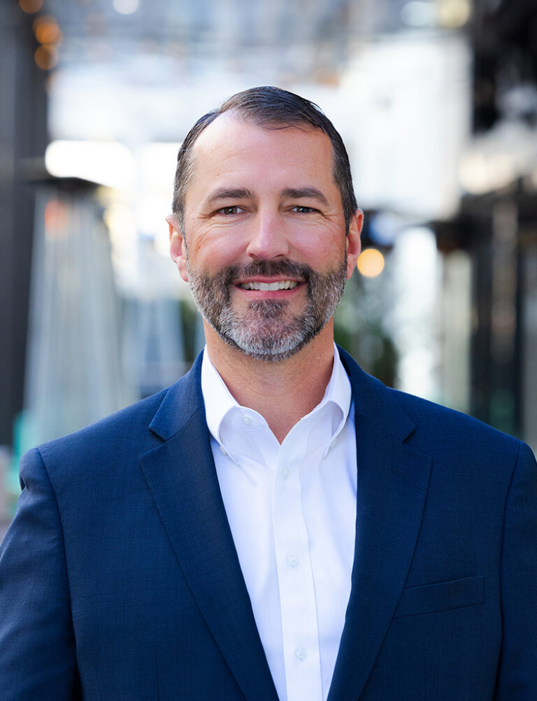 A man with short dark hair and a beard, wearing a navy blue blazer over a white button-down shirt.